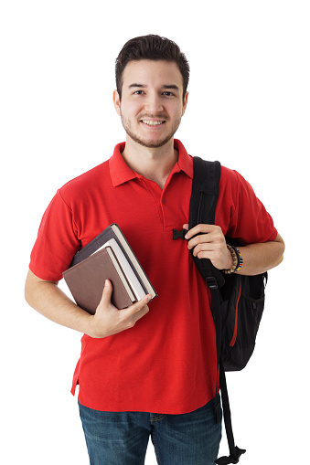 A latin male student holding a backpack and books, standing and smiling at the camera.