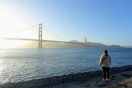 A woman admiring a beautiful view of the famous Golden Gate Bridge, on sunny evening during sunset, with a full view of the bay, in San Francisco, California, United States