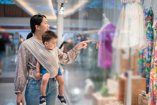 Asian woman window shopping while shopping for clothes in an unrecognizable shopping mall with her infant baby boy in a baby carrier. New mother lifestyle after having a baby