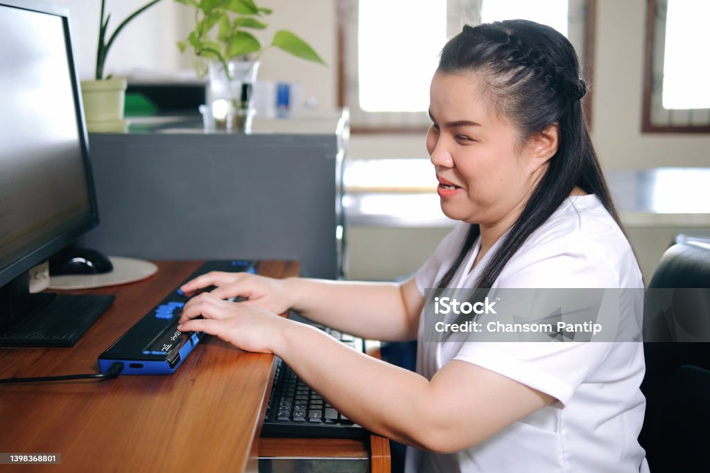 Asian woman with blindness disability using computer with refreshable braille display or braille terminal a technology assistive device for persons with visual impairment in workplace. Blindness Stock Photo