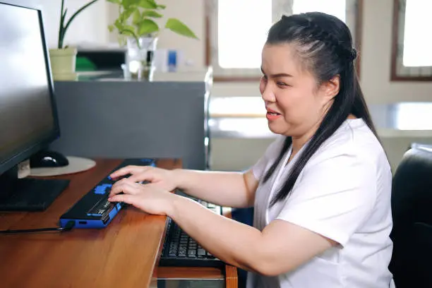 Photo of Asian woman with blindness disability using computer with refreshable braille display or braille terminal a technology assistive device for persons with visual impairment in workplace.