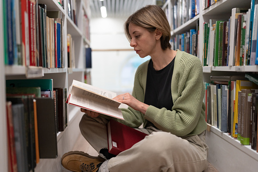 Thoughtful female mature student sits cross-legged on floor between bookshelves in library, reads book, searches for information and conduct research without internet, getting second higher education.