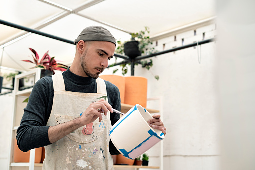 Latin American man standing in his workshop painting a flower pot with a brush while holding it with one hand. There are several flower pots on the site.