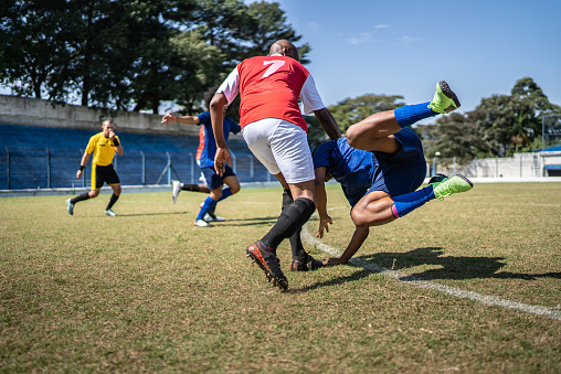 A male soccer player kicks a ball and soccer goalie jumping in motion for a ball while defending his gates on wide angle panoramic image of a outdoor soccer stadium or arena full of spectators under a sunny sky. The image has depth of field with the focus on the foreground part of the pitch. With intentional lensflares. Players are wearing unbranded soccer uniform.