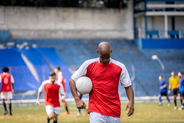 A professional male soccer player dressed in red shirt, socks and white shorts kneeling with his arms up and fists clenched, shouting in celebration having just scored a goal. The footballer is on an outdoor pitch, with other teamates and rival players in a generic football soccer stadium full of spectators.