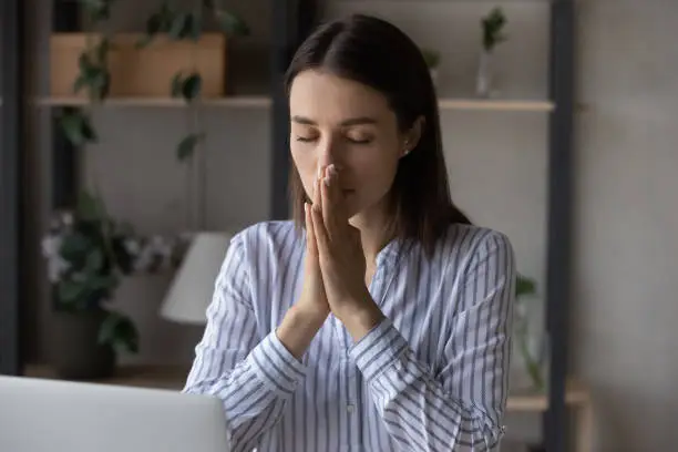 Religious millennial business woman praying at workplace. Female prayer, employee, worker asking, worshipping God, begging for help, healing, good luck, working at desk with laptop. Religion concept