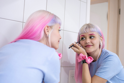 Young woman with pink hair applying make up in the bathroom mirror
