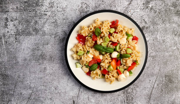 istock Pasta salad with mozzarella, greens, cucumbers and tomatoes on a round plate on a dark gray background. Top view, flat lay 1398399856