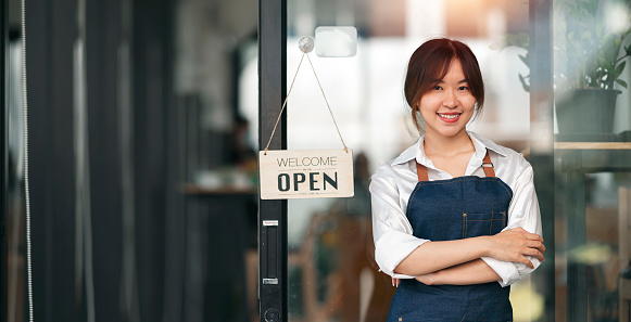 Portrait of positive asian businesswoman standing at cafeteria door entrance. Cheerful young waitress in blue jeans apron near glass door with open signboard and looking at camera.