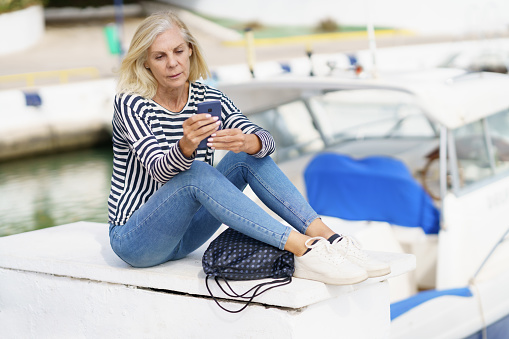 Mature woman using her smartphone sitting in a sea port. Elderly female enjoying her retirement at a seaside retreat.