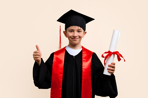 Whizz kid 9-11 year wearing graduation cap and ceremony robe shows thumb up, approval sign. Ðlementary school graduate boy with certificate diploma tied with red ribbon in studio.