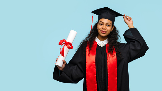 Happy Afro American Woman Graduate Wearing Ceremony Robe and Mortarboard Holding Certificate tied with Red Ribbon on Blue Background. Student Girl Celebrating Graduation and Getting Diploma