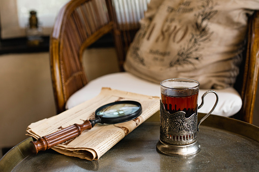 A glass in an antique cup holder with tea and old newspaper with a loupe are on a copper table against the backdrop of a Victorian-style interior.