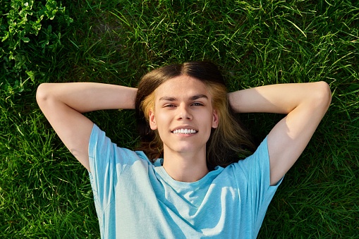 Smiling young guy looking at camera, lying on grass, green background. Handsome teenage male with long hair, trendy hairstyle in blue t-shirt. Youth, lifestyle, people concept