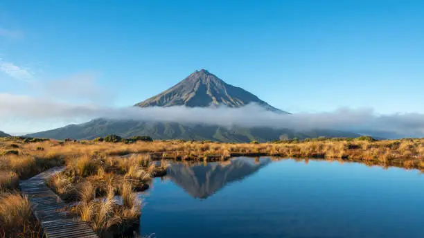 Photo of Mt Taranaki reflected in the clear water of Pouakai tarn, clouds drifting over the mountain, New Zealand.