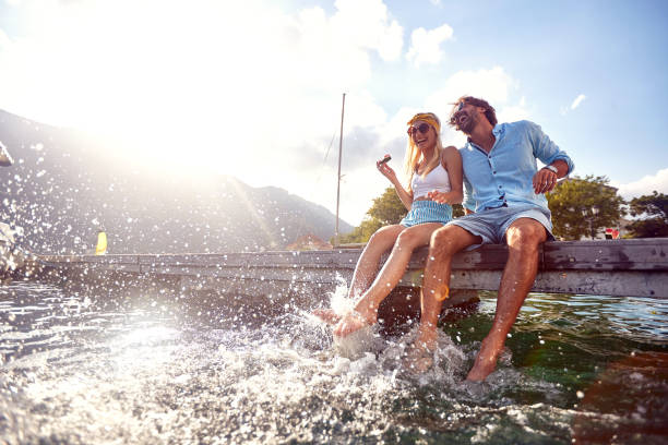 attractive couple laughing while sitting on jetty near water. splashing water with legs. couple in love. tourism, summertime, togetherness, lifestyle concept. - jetty imagens e fotografias de stock