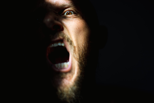 Dramatic light self portrait: bearded man in the shadow. Half of the face is illuminated by sunlight, the other is in the dark. A man plays doing a scary face with open mouth
