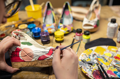 POV of a Caucasian female artist, paint the high heels with paint, while working at the shoemaker workshop