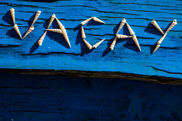 Word 'Vacay' Written With Seashells on Blue Wood From Directly Above.