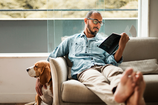 Bearded man comfortably sitting on a coach reading a book and holding his dog