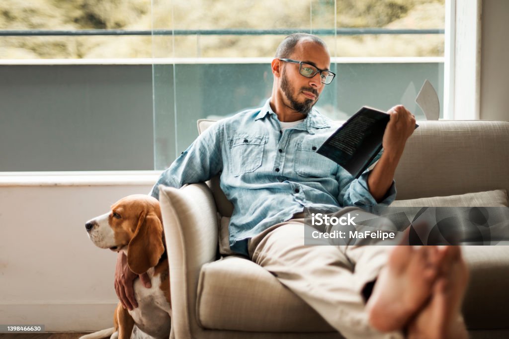 Bearded man comfortably sitting on a coach reading a book and holding his dog Bearded man comfortably sitting on a coach reading a book and holding his dog . Book Stock Photo