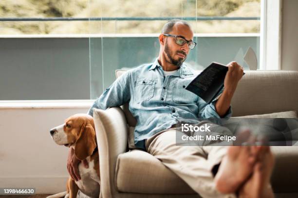 Bearded Man Comfortably Sitting On A Coach Reading A Book And Holding His Dog Stock Photo - Download Image Now
