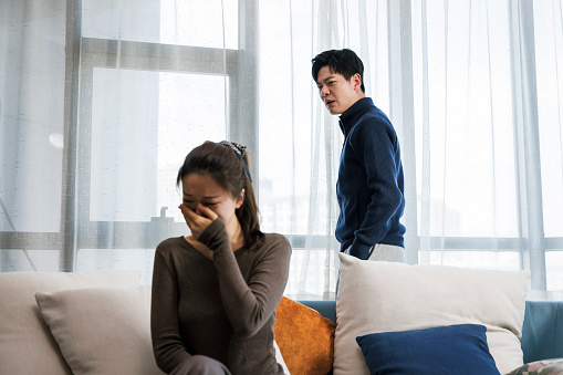 This is a young asian couple. They wear casual clothes. Sitting on the sofa in the living room at home. There was a quarrel between them, and the man was very excited. Women are sad.