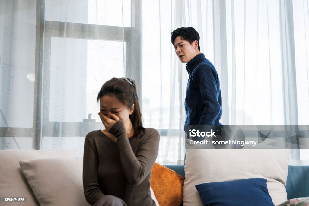 Handsome man and beautiful young woman are having quarrel. This is a young asian couple. They wear casual clothes. Sitting on the sofa in the living room at home. There was a quarrel between them, and the man was very excited. Women are sad. Arguing Stock Photo