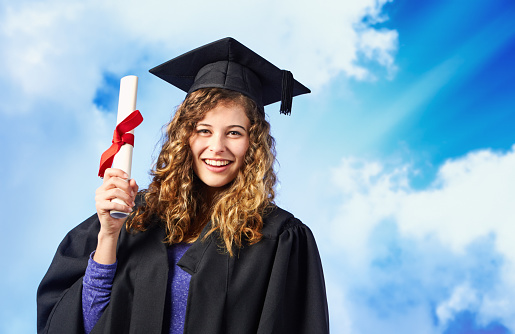 Beautiful young woman wearing mortar board and academic gown holds her diploma, tied up with a red ribbon.