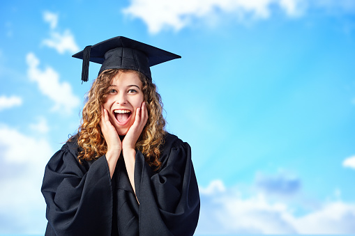 Beautiful young woman wearing mortar board and academic gown looks amazed to have graduated.