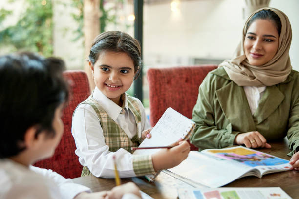 Smiling 6 year old girl in uniform sitting at dining table in Riyadh family home and practicing writing sentences in English with mother’s support.