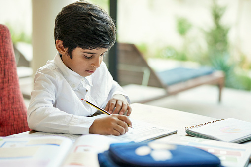 Waist-up view of 7 year old Middle Eastern boy sitting at dining table in family home and concentrating on assignment in workbook.