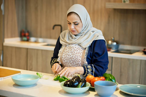 Waist-up view of Middle Eastern woman in abaya, apron, and hijab standing at counter preparing food for midday meal.