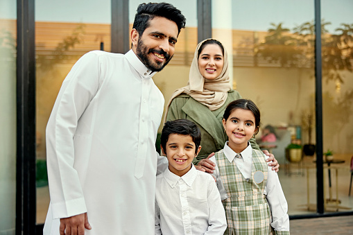 Portrait of Middle Eastern couple in traditional attire standing with elementary age siblings on deck of modern Riyadh home and smiling at camera.