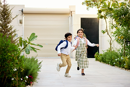Full length view of 6 and 7 year old Middle Eastern children in school uniforms holding hands and grimacing with effort as they run towards camera.