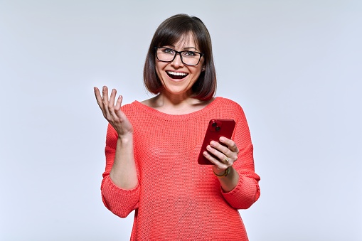 Portrait of middle aged beautiful woman with phone in hands looking at camera on light studio background. Smiling mature female in red, glasses, with make-up hairstyle. Technology, lifestyle, people
