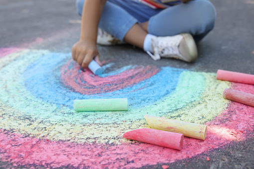 The photo of a little girl sitting on the ground and using some colorful chalk to draw flowers.