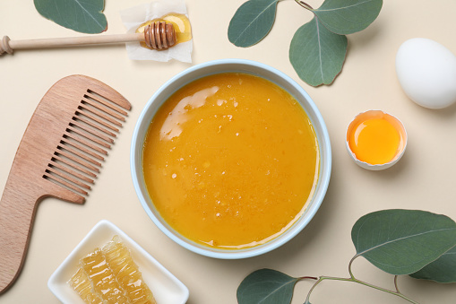 Homemade hair mask in bowl, ingredients and bamboo comb on beige background, flat lay