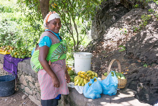 Santiago island, Cape Verde - September 03, 2015: Peasant woman with a child on her back selling fruit on a road in the Picos region in the interior of the island