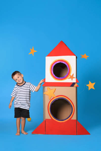istock Cute little boy playing with cardboard rocket on light blue background 1398515645