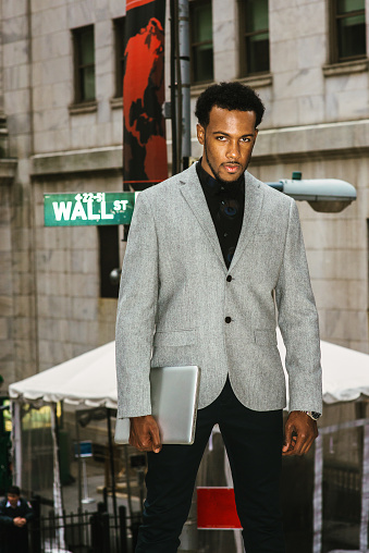 African American Businessman traveling, working in New York. Wearing gray blazer, holding laptop computer, a black man with beard standing on street, tensely looking at you.