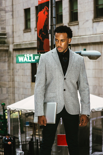 African American Businessman traveling, working in New York. Wearing gray blazer, holding laptop computer, a black man with beard standing on Wall Street, looking away. 