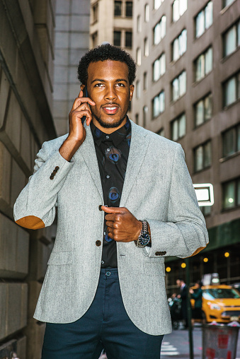 African American businessman traveling, working in New York. Young black professional with beard walking through narrow, crowded high building street, talking on cell phone.