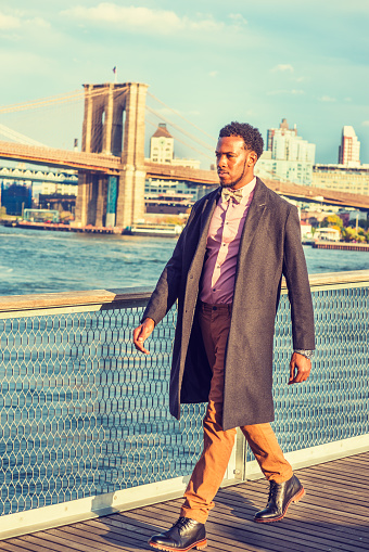 African American Businessman traveling in New York. Wearing woolen overcoat, pink shirt, brown pants, leather boot shoes, bow tie, black man with beard, confidently walking on deck at harbor in sunset. 