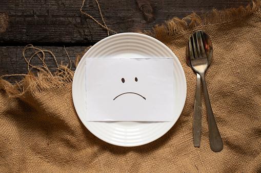 a sad smiley is drawn on paper lies on a white plate, on a wooden table and on a brown tablecloth, sadness and depression, crisis has nothing to eat