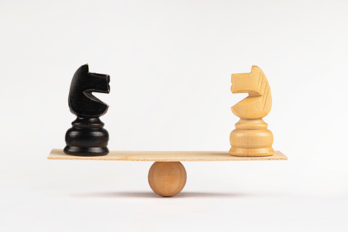 White and a black chess knight horses stand on wooden seesaw on white background