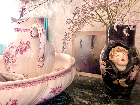 Horizontal closeup photo of an ornate cream ceramic wash basin and jug decorated with pink floral designs, on a marble shelf, next to a black retro style vase with dried Statice flowers and decorated with a child’s face and flowers. The Camargue, Provence, France.