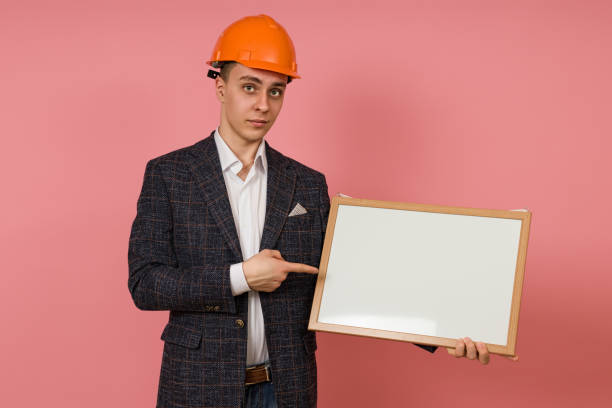 istock young engineer in a protective helmet and jacket points with a finger at a white board in his hand on a pink background 1398550773