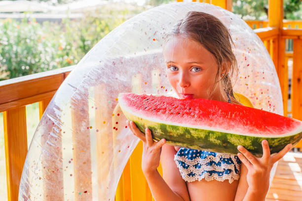육즙이 많은 수박을 먹는 공원의 귀여운 소녀. 여름 테라스에있는 소녀는 붉은 수박의 큰 조각을 먹고 미소를 짓습니다. 음식 자연. 사랑스러운 금발 소녀는 야외에서 수박 조각을 먹습니다. - watermelon missing bite melon grass 뉴스 사진 이미지