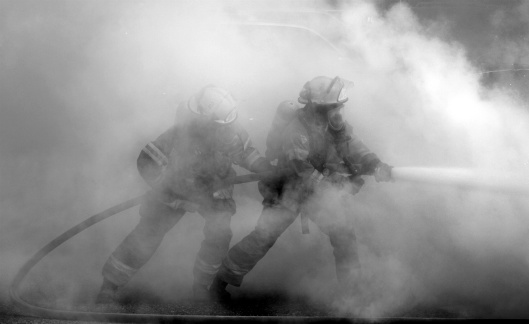 Two firemen engulfed in smoke fighting a carfire with a waterhose.